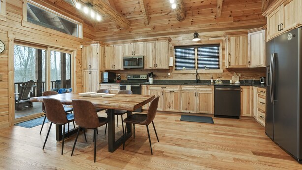 A wooden cabin interior with a dining area, kitchen, and large windows.