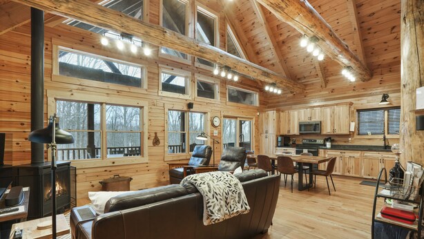 A cozy cabin interior with a fireplace, leather sofa, dining area, kitchen, and large windows. - Falling Leaves | Hocking Hills, Accessible, Pets (South Bloomingville)