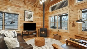 A cozy wooden cabin interior with a fireplace, leather sofa, wooden tables, and a television.