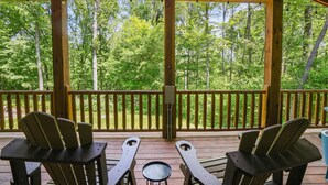 A wooden deck with Adirondack chairs and a small table, overlooking a forested area.