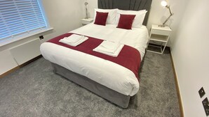 Una cama bien hecha con ropa de cama blanca y roja, dos mesitas de noche con lámparas y cuadros en las paredes.