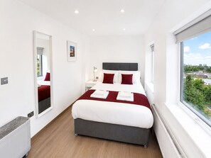 A modern hotel room with a large bed, a bedside table, a mirror, and a window with a city view. - Roomspace Apartments - Harvest Lane (Thames Ditton)