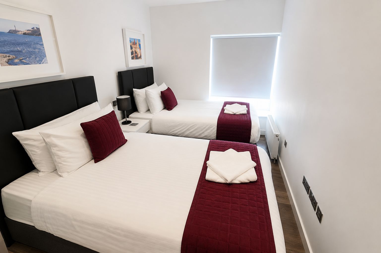 A hotel room with two beds, a nightstand, a lamp, and a window with blinds.