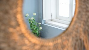 A view through a wooden-framed mirror reflecting a window with white shutters, a white wall, and a potted plant with white flowers. - GuestReady - Souto Downtown Studio 1 (Porto)