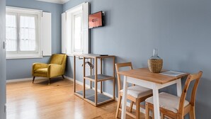 A room with a wooden floor, a yellow armchair, a wooden table with chairs, a wall-mounted TV, and a window with white curtains. - GuestReady - Souto Studio 4|4th floor|No elevator (Porto)