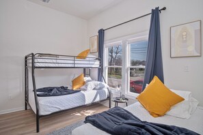 A room with a bunk bed, a single bed, a small table, and a window with curtains.