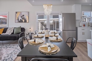 Un comedor moderno con una mesa de madera oscura dispuesta para seis personas, rodeada de sillas negras. Un candelabro cuelga encima, y un amplio ventanal ofrece luz natural.