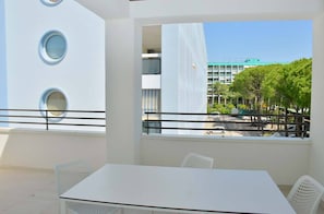 A modern outdoor seating area with a white table and chairs, a balcony with a railing, and a view of a building and trees in the background.