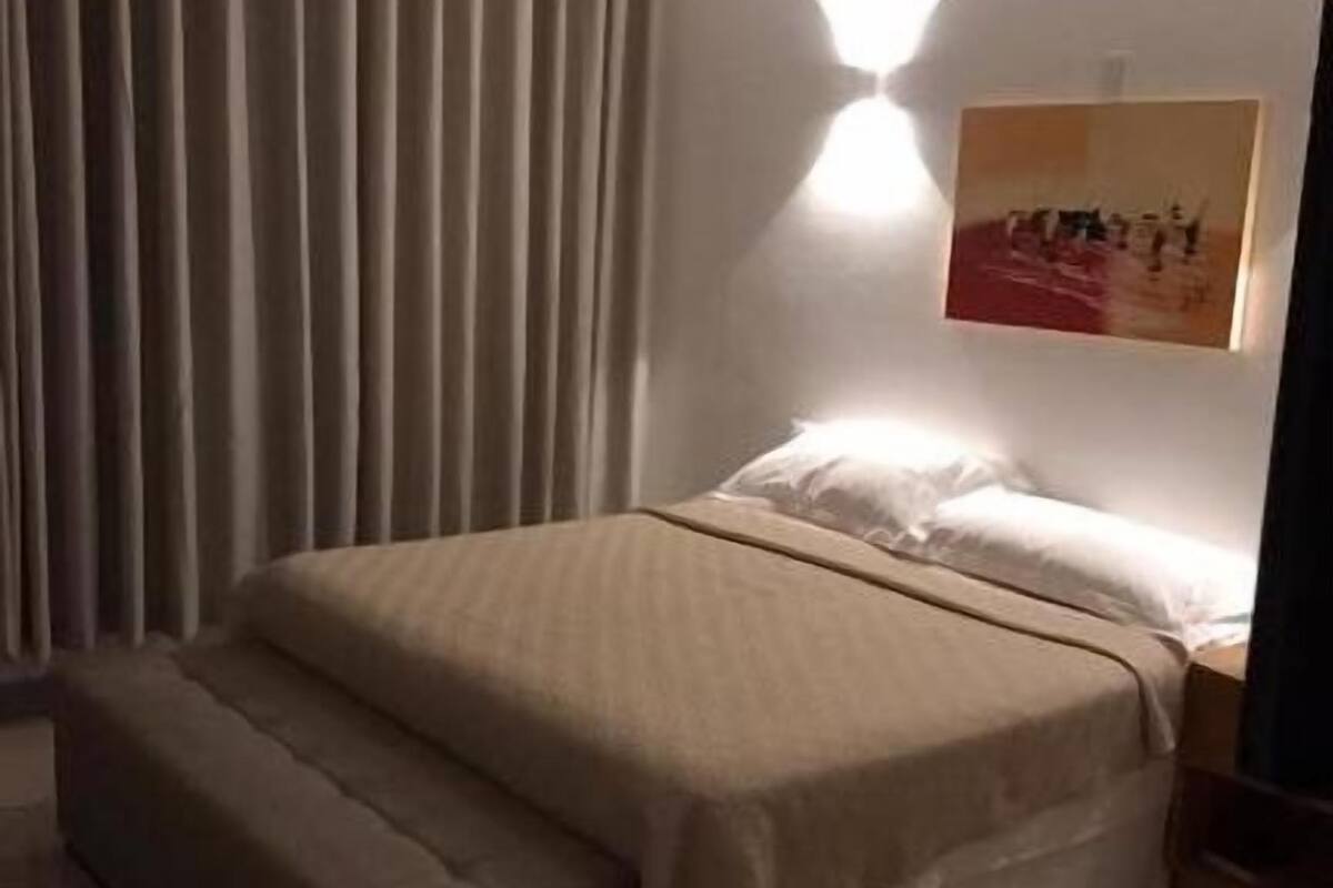 A hotel room with a bed, a bench, a wall-mounted light, and a painting.