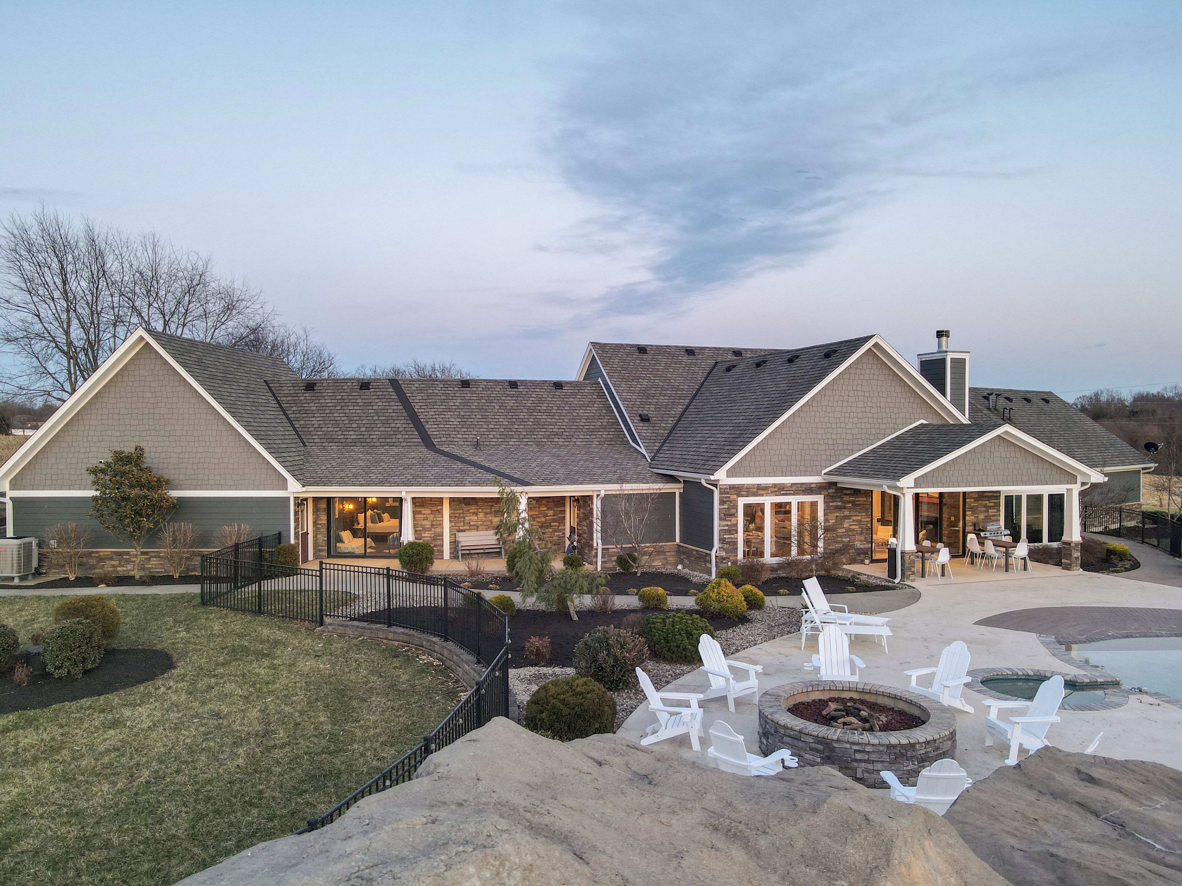 A two-story house with a stone facade, a covered patio with Adirondack chairs, and a fire pit.