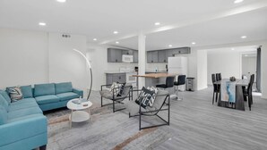 A modern living room with a blue sofa, a coffee table, and a floor lamp. There is a kitchen area with a dining table and chairs.