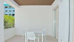 Un balcón con mesa y sillas blancas, un ventanal de vidrio y vistas a un edificio y árboles.