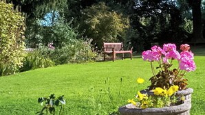 A garden with a stone planter containing flowers, a wooden bench, and a grassy area. - 2 bedroom accommodation in Weobley, Herefordshire (Weobley, Herefordshire)
