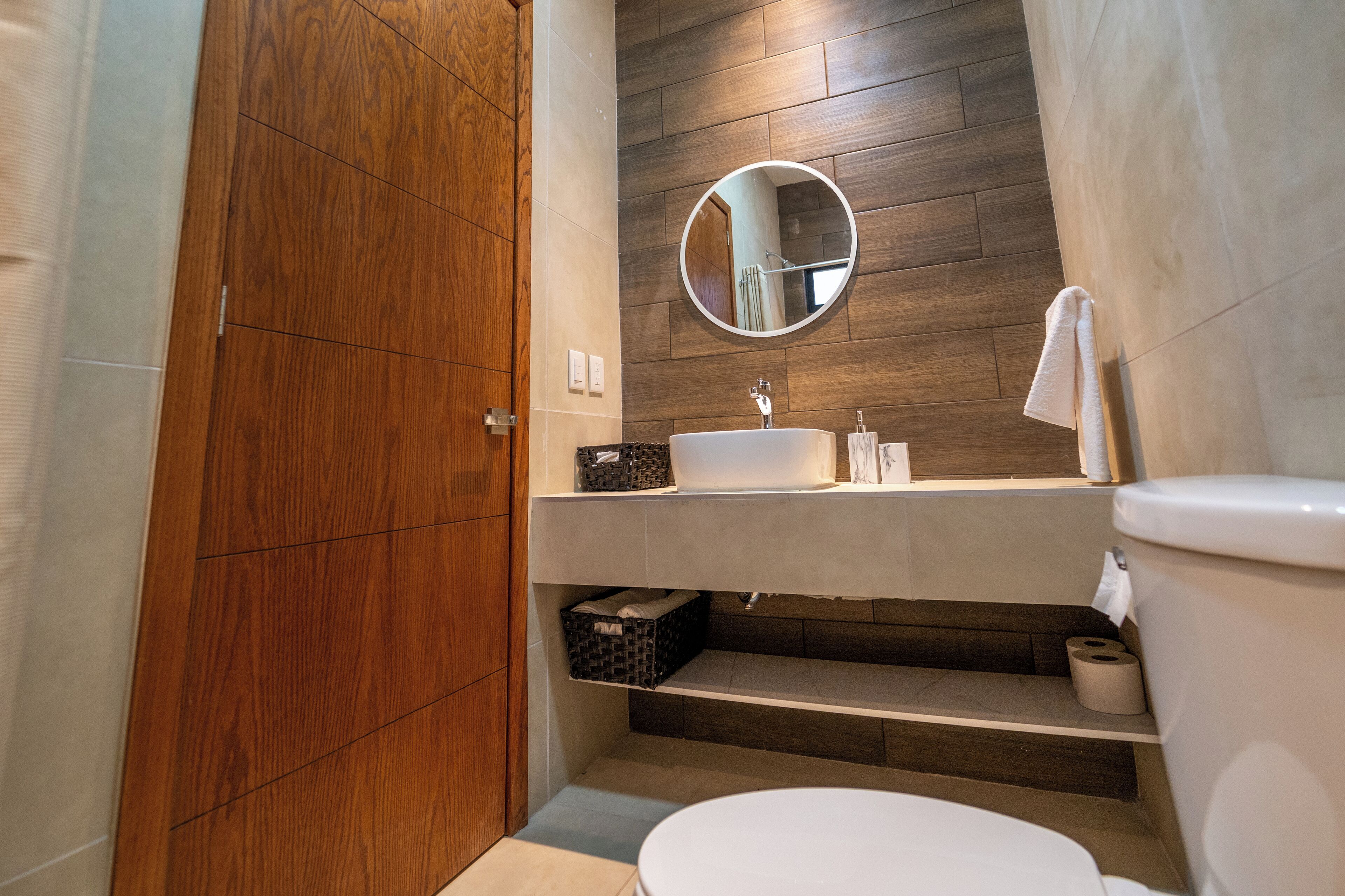A modern bathroom with a wooden door, a round mirror, a white sink, and a toilet.