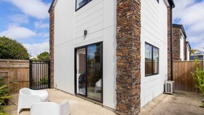 A modern house with a stone facade, large sliding glass doors, and a covered patio area with white chairs.