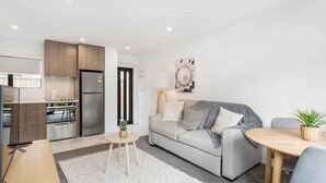 Una sala de estar moderna con un sofá gris, una mesita redonda y una zona de cocina con nevera y estufa.