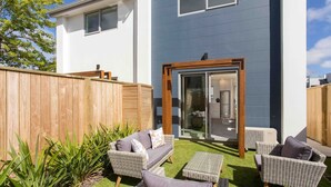 Una casa moderna con fachada azul, cerca de madera y un patio con mobiliario de mimbre y un pequeño jardín.