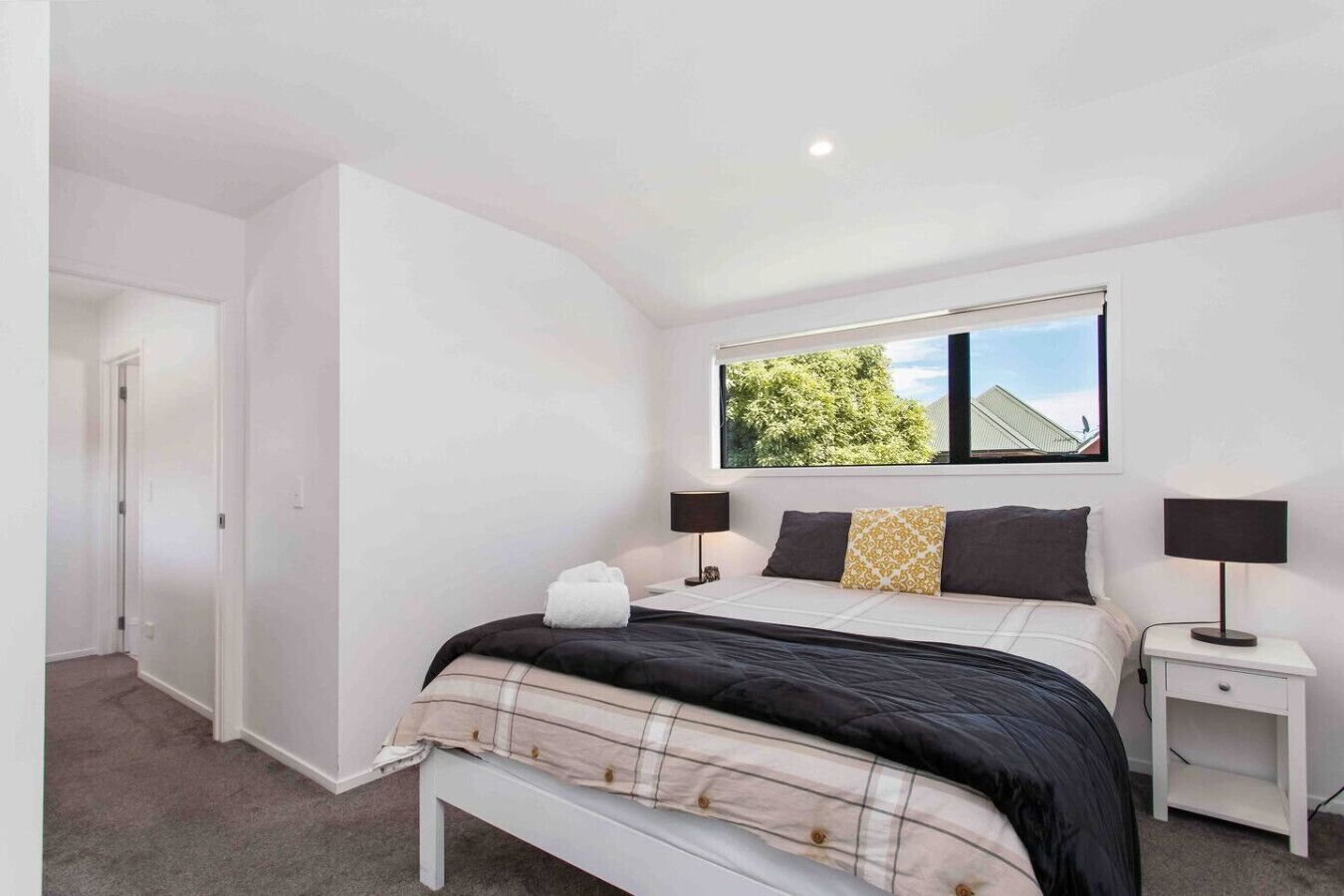 A bedroom with a bed, two bedside tables, a window with a view of trees, and a door leading to another room.