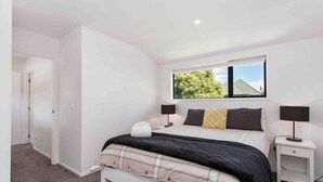 A bedroom with a bed, two bedside tables, a window with a view of trees, and a door leading to another room. - Relax Unwind Super Fast Internet (Christchurch)