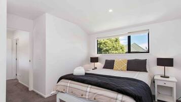 A bedroom with a bed, two bedside tables, a window with a view of trees, and a door leading to another room.