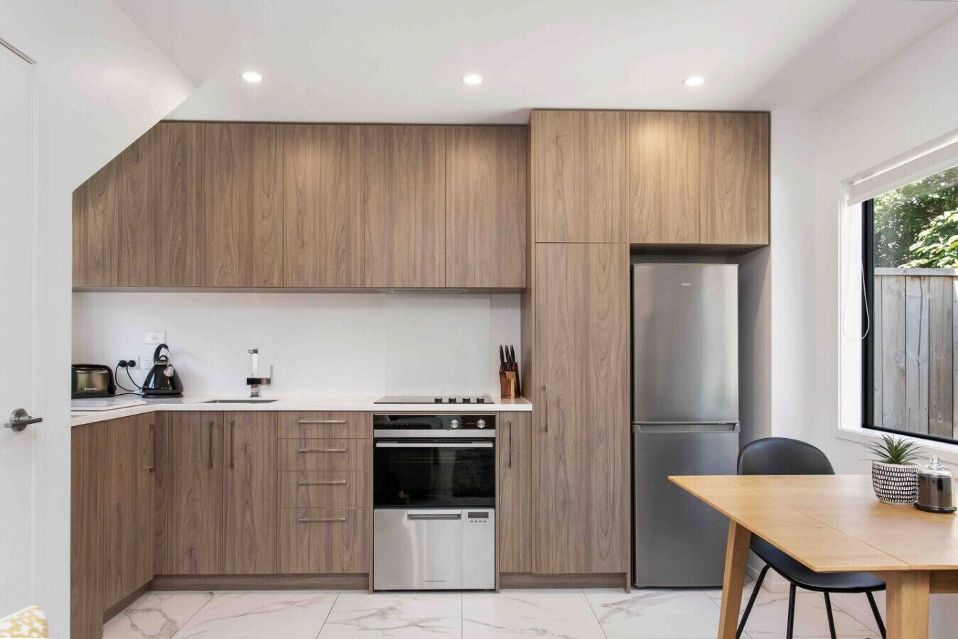 A modern kitchen with wooden cabinets, a stainless steel refrigerator, a built-in oven, and a dining area with a table and chairs.