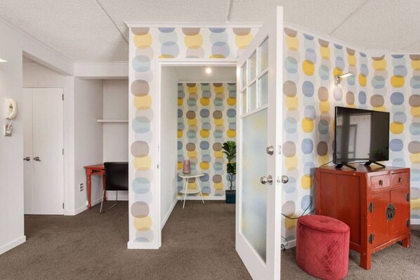 Una habitación con papel tapiz de lunares, un televisor sobre un mueble de madera, una mesita con una planta y un otoman rojo.