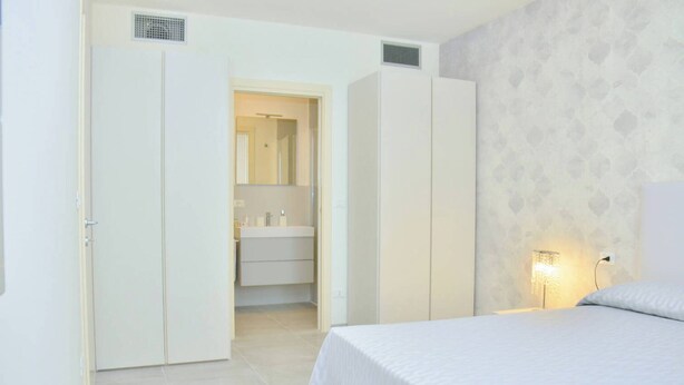 A bedroom with a bed, a nightstand, a wall-mounted lamp, and a bathroom with a sink and mirror. - Relax Vibes Near the Beach (San Michele al Tagliamento)