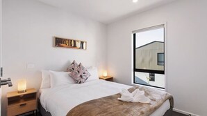 A modern bedroom with a large bed, a nightstand with a lamp, a painting on the wall, and a window with a view of a building.