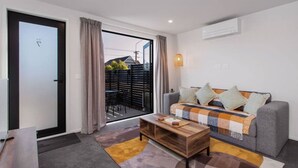 A modern living room with a grey sofa, a wooden coffee table, and a view through a sliding glass door to an outdoor area with a black fence.