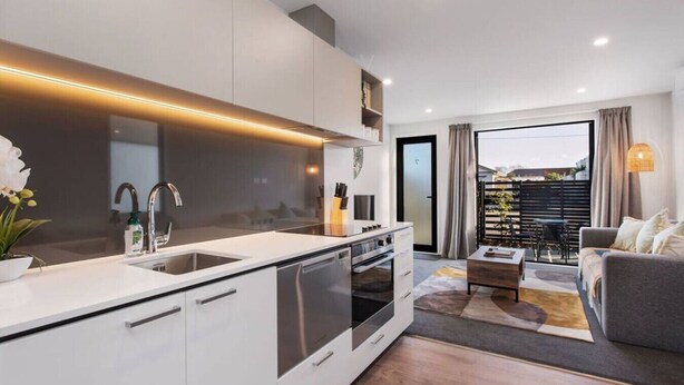 A modern kitchen with a central island, built-in oven, and stainless steel appliances. There is a dining area with a grey sofa and a coffee table.