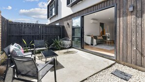 A modern house with a patio area featuring outdoor furniture, a wooden fence, and a sliding glass door leading to an interior space with a sofa and a dining table.