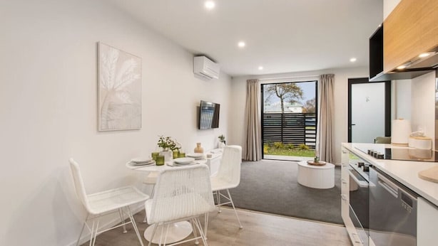 A modern kitchen with white furniture, a dining area with a table and chairs, and a large window offering a view of the outdoors. - Style Location New Sunny 2Bdrms Free Park (Christchurch)
