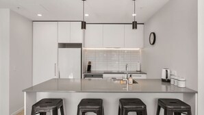 A modern kitchen with a central island, three stools, a refrigerator, a sink, and a clock on the wall.