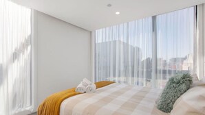 A modern hotel room with a large bed, a view of the city through sheer curtains, and a cityscape visible through the window.