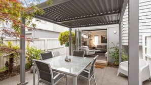 Une terrasse avec une table et des chaises en verre, un espace salon couvert et une vue sur l’intérieur de la maison.
