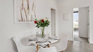 Un comedor moderno con una mesa blanca preparada para cuatro, un jarrón con flores rojas y blancas, y una obra de arte enmarcada con hierbas altas en la pared.