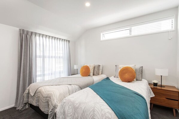 A modern hotel room with two beds, a large window with blinds, a nightstand with a lamp, and a view of the city.