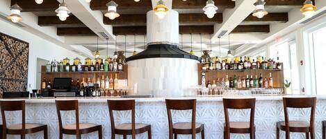 A bar area with a high ceiling, wooden beams, and hanging pendant lights. There are bar stools and a counter with a variety of bottles and glasses.