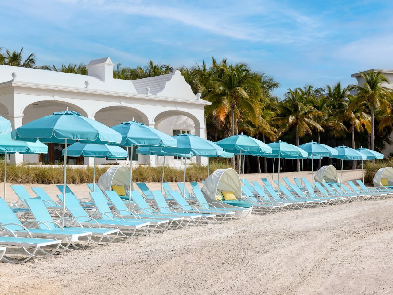 A row of blue sun loungers with white canopies, lined up on a sandy beach in front of a white building with arches and palm trees in the background.