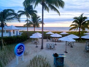Un restaurante de sushi frente a la playa con mesas al aire libre, sombrillas, palmeras y vistas al océano.