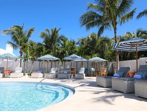 Un área junto a la piscina con sillas de descanso, sombrillas y palmeras.