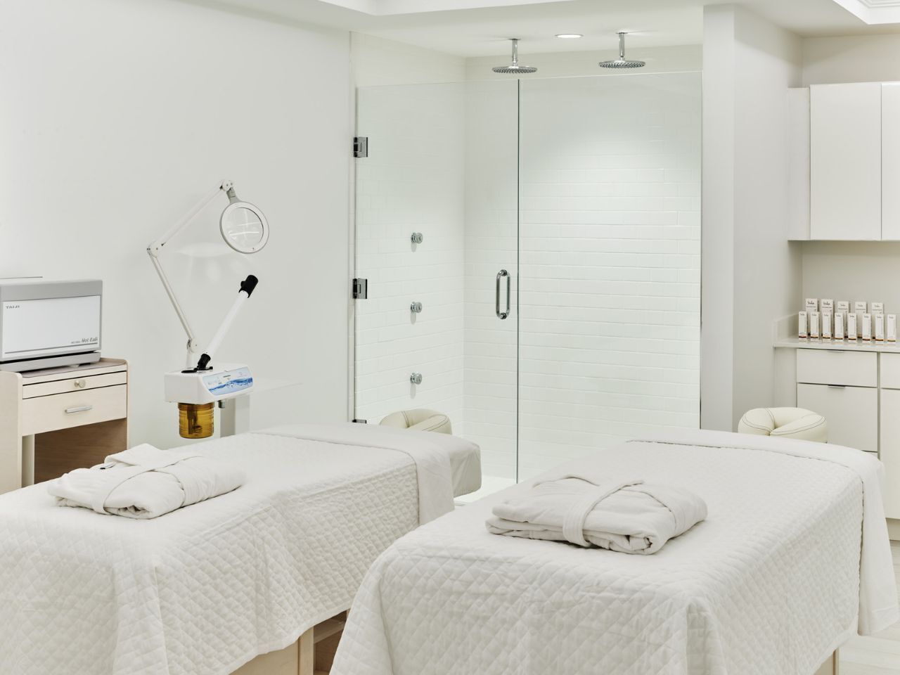 A modern hotel room with two beds, a desk, a computer, a massage chair, and a shower area.