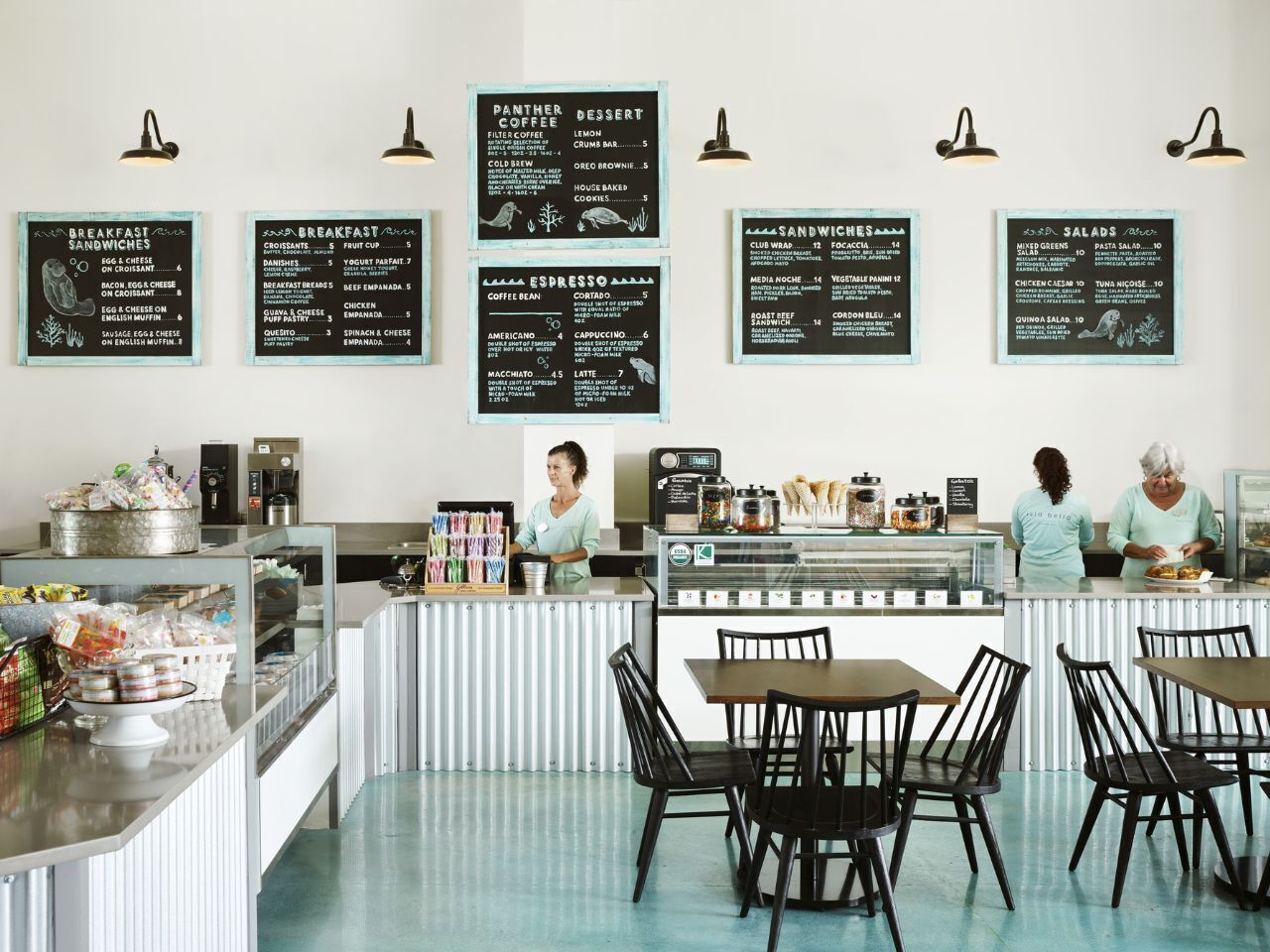 Una cafetería con varios menús que ofrecen desayuno, sándwiches, café espresso y postres. Hay mesas y sillas para sentarse, y una barra con una máquina de café y alimentos.