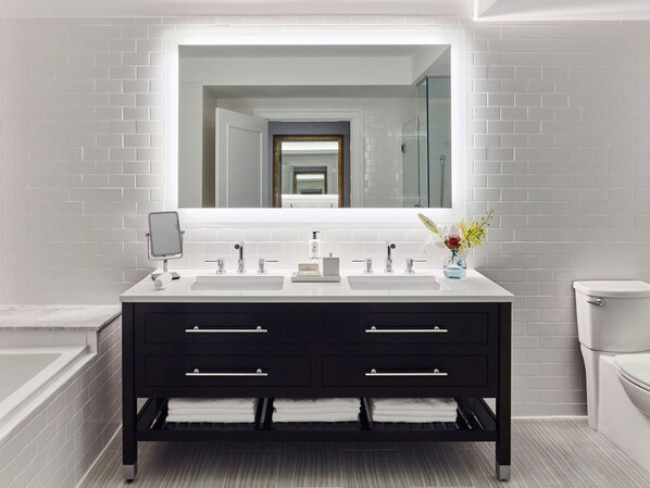 Une salle de bain moderne avec un mur recouvert de carreaux blancs, un meuble-lavabo noir doté de deux vasques, un miroir et des toilettes.
