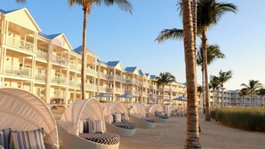 Un hôtel en bord de mer, composé de bâtiments à plusieurs étages, doté d’espaces extérieurs aménagés avec des parasols et bordé de palmiers.