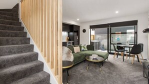 A modern living room with a staircase, a sofa, a dining table, and a kitchen in the background.