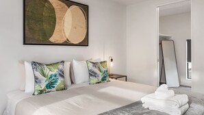 A modern hotel room with a large bed, two pillows with tropical leaf patterns, a framed abstract painting, a bedside lamp, a full-length mirror, and neatly folded towels on a marble table.