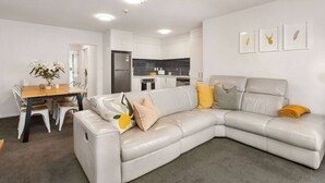 A modern living room with a grey sectional sofa, a dining table with chairs, a kitchen area, and a small dining table with flowers.