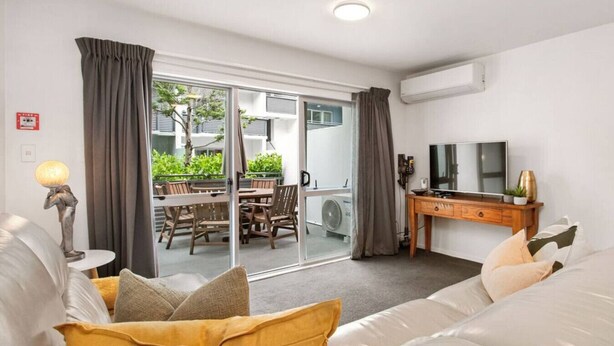 A modern hotel room with a sofa, a TV, a wooden side table, and a sliding glass door leading to an outdoor area with a table and chairs.