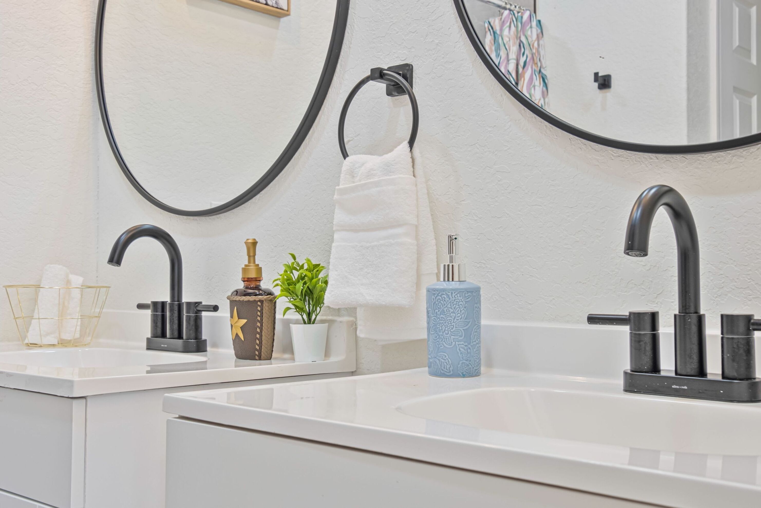 A modern bathroom with two sinks, black faucets, a white towel, a small potted plant, and a blue soap dispenser.